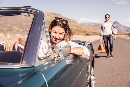 Woman in convertible car on a road trip waiting for man with a travelling bag