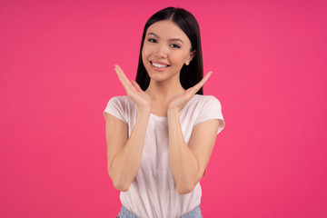Obraz na płótnie Canvas Pleasant looking Asian female model smiles gladfully, spreads palms near face, expresses positive emotions, wears white t shirt, has appealing appearance, healthy skin, isolated on pink studio wall