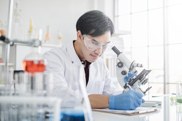 A male scientist with black hair wearing white coat and protective glassware writing and looking into a microscope in a laboratory setting with test tubes.