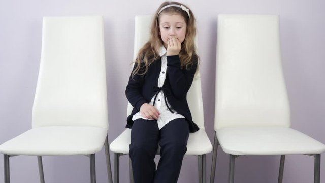 girl in elementary school is worried, bites her nails, thinks on a white chair, worries.