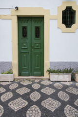 Portugal: Alte grüne Haustür und Fenster umrahmt von Natursteinen mit davor geometrisch verlegten Pflastersteinen 