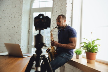 African-american musician playing saxophone during online concert at home isolated and quarantined. Using camera, laptop, streaming, recording courses. Concept of art, support, music, hobby, education