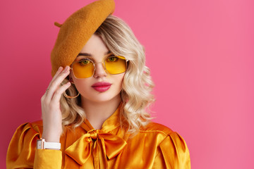 Fashionable woman wearing yellow sunglasses, beret, silk blouse, white wrist watch, posing on pink background. Copy, empty space for text