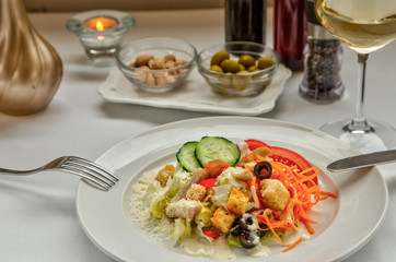 Delicious fresh Caesar salad with croutons, bacon, lettuce, vegetables, parmesan and mayonnaise. Rosettes with almonds and green olives and a glass of white wine on the table