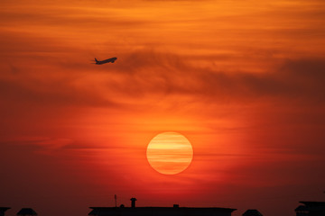 Tiny airplane silhouette flying on orange sunset sky with big sun disk