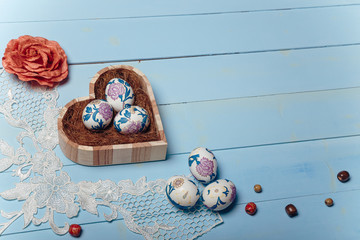 lose up shot of Easter eggs in wooden heart shaped box with decorative elements on blue background. Easter and spring concept. Top view, copy space.