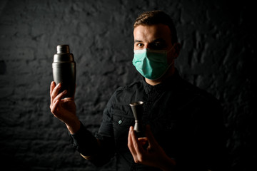 man in mask stands in dark bar and holds shaker and jigger in his hands