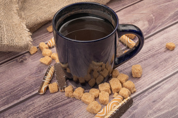 Dark blue ceramic mug with tea, cookie sticks with chocolate and white icing and pieces of brown cane sugar on a wooden background. Close up.