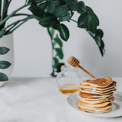 Pancakes on a white plate. Behind them is a jar of honey and a spoon. Against the background of a flower Zamioculcas in a white flower pot.
