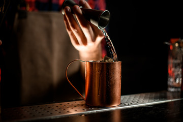 close-up bartender's hand pours drink from jigger into steel mug full of ice cubes with condensate