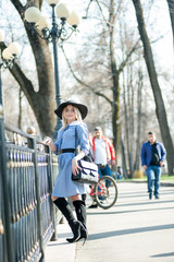 young pretty blond woman in blue dress and black hat posing on park on sunny day background