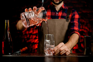 professional bartender accurate pours cocktail from mixing cup into small glass.