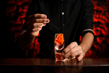 male bartender holds tweezers over glass with drink and slice of citrus