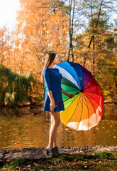 Beautiful young girl with long hair in a blue short dress with a rainbow umbrella walks through the autumn park.