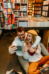 Young student couple surrounded by books is taking selfie in the library while sitting on the floor.