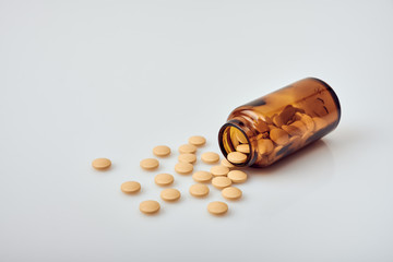 Yellow round tablets scattered from a small brown glass bottle without lid laying on white acrylic table