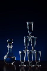 Crystal decanter or carafe of vodka and six glasses placed as pyramid tower on wooden table in restaurant, blue and black background with copy space