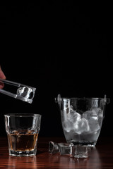 hand with tongs holding ice-cube above a glass with whiskey, ice bucket behind the alcoholic drink on wooden table in restaurant. Black background for copy space