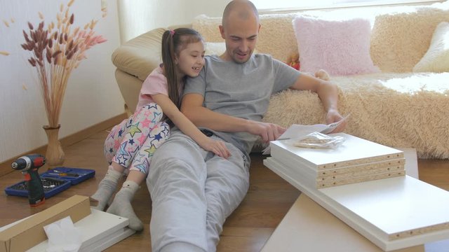A father with his beautiful little daughter opens a package with new furniture to start her assembling. The concept of a happy family and good relationships. Raising children and initiation to work.