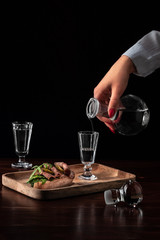 female hand holds crystal decanter or carafe and pours on vodka in a glass. tasty appetizer bruschetta with bacon and pickles on wooden board. alcohol drink concept with copy space