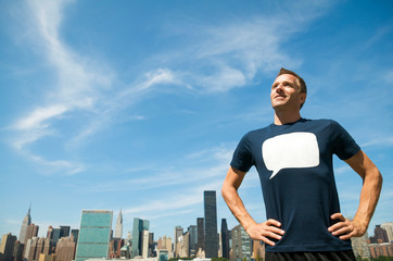 Confident young man standing outdoors in a T-shirt with a blank speech bubble above the city skyline under sunny blue sky