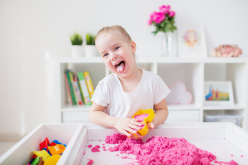 Little blonde girl playing with pink kinetic sand on a white table in a light room. Sensory...