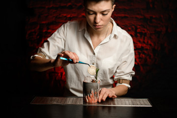 Woman bartender accurate decorates figured glass with ice by slice of citrus using tweezers