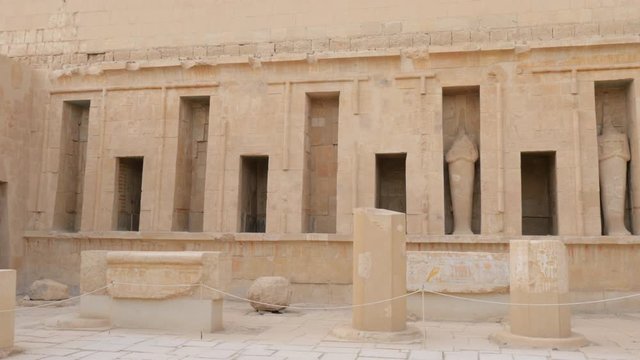 There Are Niches in the Wall with Destroyed Statues. Ruined Parts of Columns are Near the Wall. Ancient Inscriptions are on the Columns.