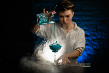 Woman bartender pours blue cocktail from mixing cup into glass at smoky bar