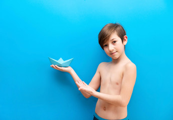 A boy in swimming trunks holds a paper boat on a blue background. Background travelers, sea adventures. Fun at the water park and pool