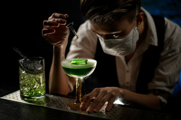 Woman bartender in mask carefully add ingredient to smoky glass with green cocktail.