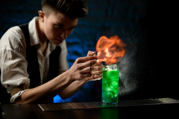 bartender sprinkles on glass with bright green cocktail and makes fire flame over it.