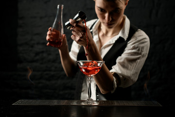 Young female bartender professionally pours drink into glass which standing on burning bar counter