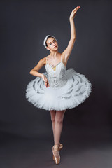 Fototapeta premium A young graceful ballerina dressed in professional attire, pointe shoes and a white tutu, demonstrates dance skills. Beautiful classic ballet dancer isolated on grey backgroung in photostudio