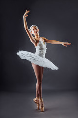 Portrait of the ballerina in the role of a white swan on grey background. 
Beautiful, attractive, young, graceful girl professionally performing ballet pas, dressed in a swan costume and pointe shoes