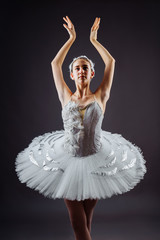 Portrait of the ballerina in the role of a white swan on grey background. Beautiful, attractive, young, graceful girl professionally performing ballet pas, dressed in a swan costume and pointe shoes