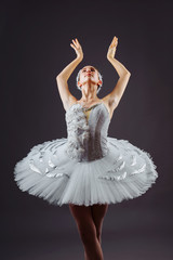 Fototapeta na wymiar Portrait of the ballerina in the role of a white swan on grey background. Beautiful, attractive, young, graceful girl professionally performing ballet pas, dressed in a swan costume and pointe shoes