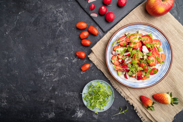 Vegetarian fruits and vegetables salad of strawberry, kiwi, tomatoes, microgreen sprouts on black concrete background. Top view, copy space.