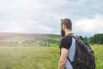 Handsome man hipster with beard on serious face sunny outdoor on mountain top against cloudy sky on natural. Tourism concept. Travel with a backpack. Wonderful landscape.