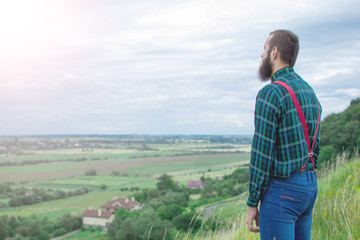 Naklejka na ściany i meble Handsome man hipster with beard on serious face in cloth shirt and suspenders sunny outdoor on mountain top against cloudy sky on natural. Tourism concept. Wonderful landscape.