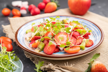 Vegetarian fruits and vegetables salad of strawberry, kiwi, tomatoes, microgreen sprouts on black concrete background. Side view, selective focus.