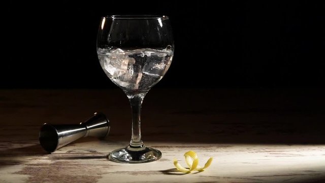 Person pouring ice cubes into glass of gin tonic with lemon and jigger on table in dark room