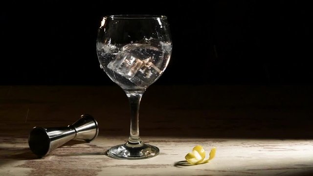 Person pouring ice cubes into glass of gin tonic with lemon and jigger on table in dark room