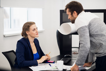 Two cheerful colleagues chatting in the office. The girl is sitting and the guy is leaning forward while standing at her desk