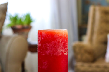 A red pillar candle in focus in the foreground with a chair and plant out of focus in the background 