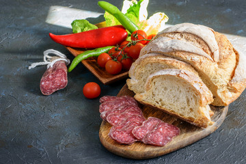 Fresh fuet and sliced homemade bread on the wooden cutting board, fresh vegetables on the dark background.