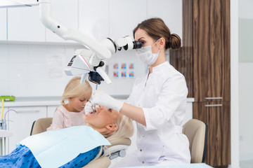 A dentist looking into a woman's mouth through a dental microscope, the woman's daughter is beside her mom, she is bored, waiting for her to complete the check up