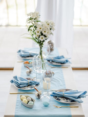 Decorative festive table setting with sky blue textile napkin and tablecloth, eggs, floral dish and golden cutlery, glass for wine. Happy easter concept.