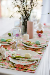 Decorative festive table setting with floral tablecloth, egg, dish and golden cutlery, glass for wine, willow branch. Happy easter holidsy concept.