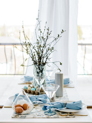 Decorative festive table setting with sky blue textile napkin and floral tablecloth, easter eggs, dishes and golden cutlery, glasses. willow in vase. holiday home interior.